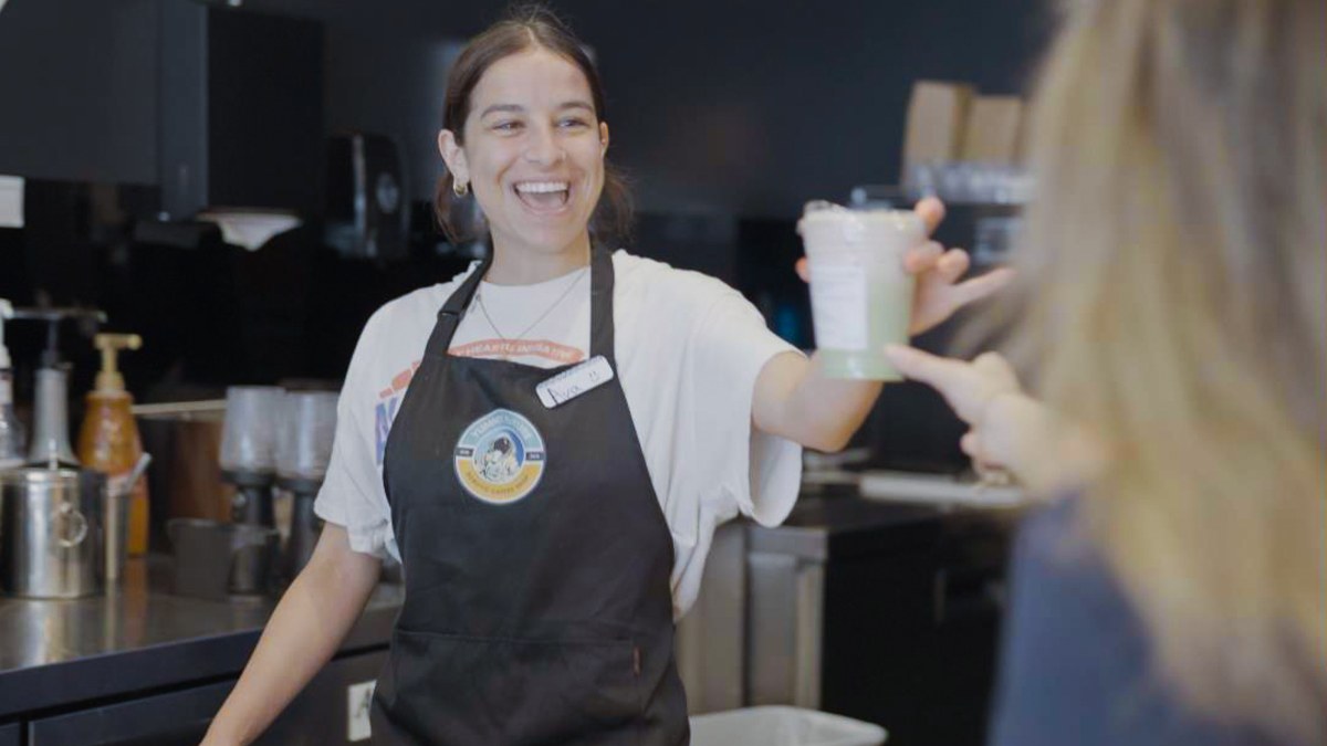A barista laughing while handing a matcha cup across the counter to a customer